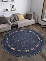 A modern living room with the Wanderer's Way - Washable Rug - JR5101 in blue, featuring colorful geometric patterns, a gray sectional sofa, assorted cushions, a round table, and wall shelves—ideal for high-traffic spaces.
