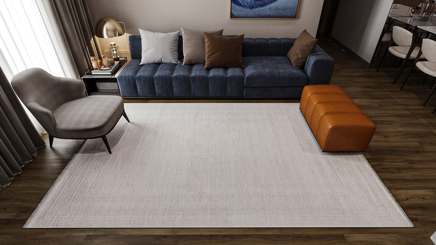 A modern living room features a blue sofa, beige and brown pillows, a gray armchair, brown leather ottoman, dark wood flooring, abstract wall art, and the Vanilla Dream Modern Bordered Rug - EW2742 in light neutral tones.