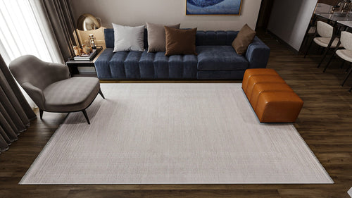 Load image 1 in gallery view. A modern living room features a blue sofa, beige and brown pillows, a gray armchair, brown leather ottoman, dark wood flooring, abstract wall art, and the Vanilla Dream Modern Bordered Rug - EW2742 in light neutral tones.