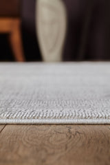 Close-up side view of the Vanilla Dream Modern Bordered Rug - EW2742 in textured light gray, lying flat on a wooden floor with blurred furniture in the background.
