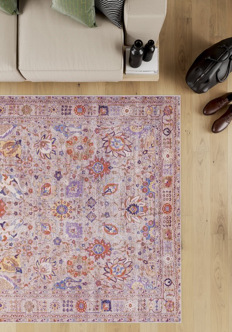 The Velorio Floral Washable Rug - LCC3006, featuring pink, purple, and orange motifs, lies on a light wood floor beside a beige sofa, black bag, brown shoes, and books—ideal for high-traffic and allergy-sensitive homes.