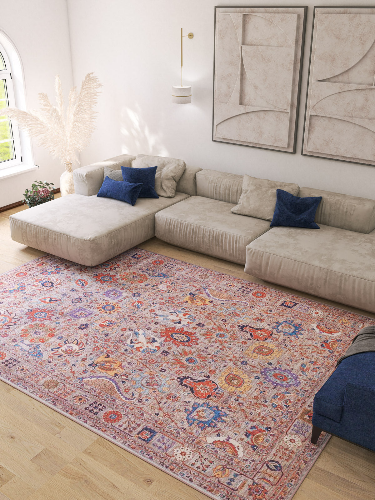 A modern living room with a light beige sectional, blue pillows, neutral wall art, pampas grass in a vase near a large arched window, and the allergy-friendly Velorio Floral Washable Rug - LCC3006 (Custom Size)—ideal for high-traffic areas.
