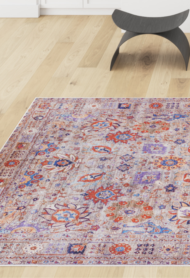 The Velorio Floral Washable Rug - LCC3006 (Custom Size), featuring intricate red, blue, and purple patterns, lies on a light wooden floor near a modern black curved stool—ideal for allergy-sensitive or high-traffic bright rooms.