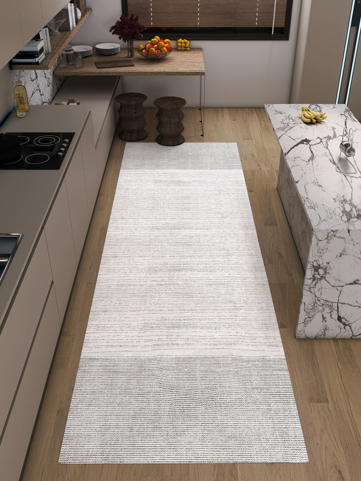 Modern kitchen with light wood floors, neutral cabinets, a marble island, and the Velvet Echo - Washable Rug - JRN25 in a gradient grey and white design—ideal for high-traffic spaces. A fruit bowl rests on a wooden table with two stools below.