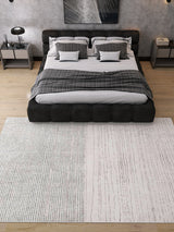 A modern bedroom with a large bed featuring gray and plaid bedding, two matching nightstands with lamps, and the Velvet Echo - Washable Rug - JRN25 (Custom Size) ideal for high-traffic areas on a light wood floor. Geometric art hangs above the bed.