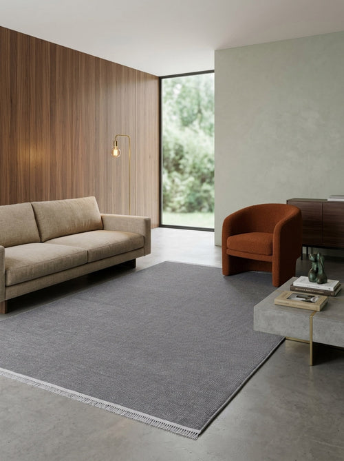 Load image 1 in gallery view. A modern living room with a beige sofa, burnt orange armchair, minimalist style, Velvet Touch Dark Gray Area Rug - PST004, concrete coffee table with books and decor, wood-paneled and green accent walls, and a large window overlooking greenery.