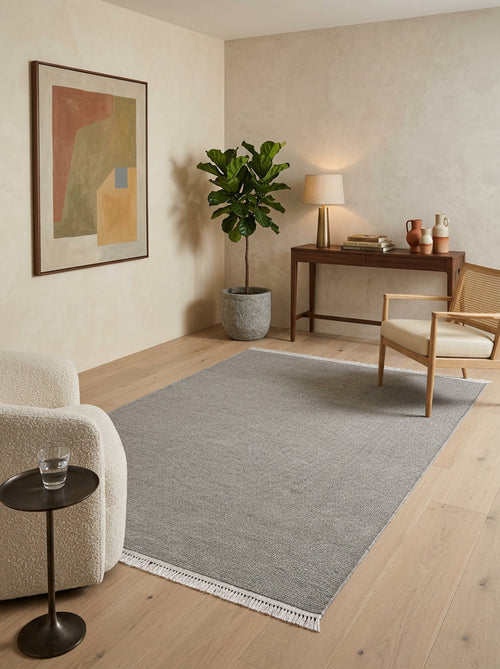 Bild 1 in Galerieansicht laden. A minimalist living room with light wood floors, a cozy white chair, small side table, wooden desk, woven chair, potted plant, table lamp, abstract wall art, and the Velvet Touch Gray Area Rug - PST003 for a soft-touch feel.