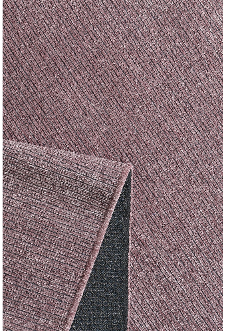 A close-up of the Velvet Touch Pink Area Rug - PST004 shows its textured, mauve surface with diagonal lines. A folded corner reveals a dark, non-slip backing—perfect for adding contemporary style to any space.