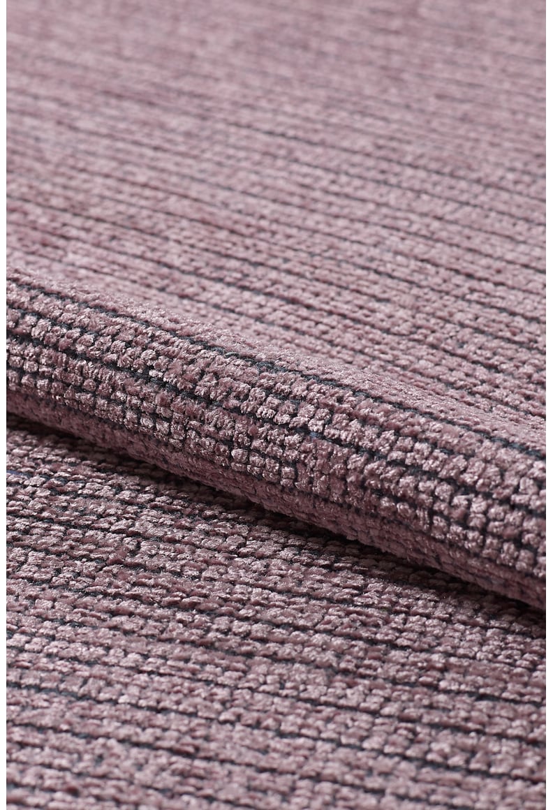 A close-up of the Velvet Touch Pink Area Rug - PST005 shows its mauve textured weave and subtle geometric pattern. Partially rolled, it highlights thick, soft material—ideal for bringing contemporary style to any space.