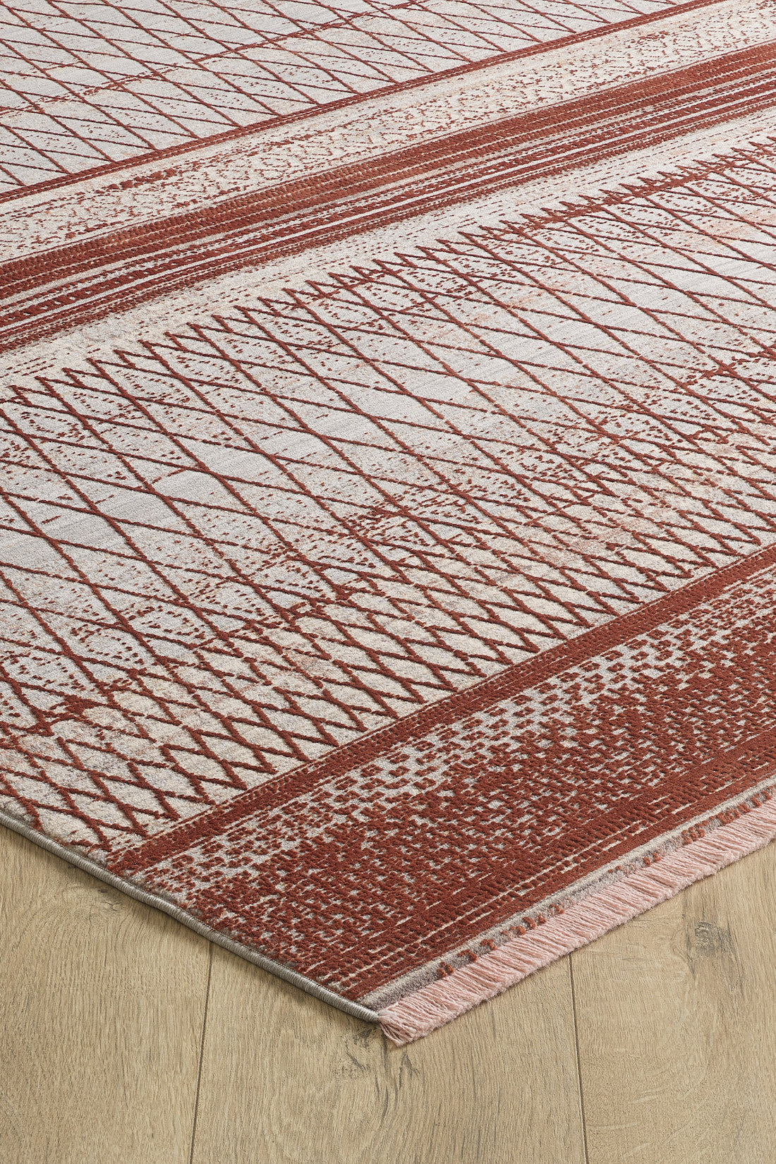 A close-up of the Vibrant Bohemian Trellis Rug - HRD009, a machine-made viscose-polyester rug with rust red and off-white geometric patterns, including diamonds and stripes, and fringed edges on a light wood floor.