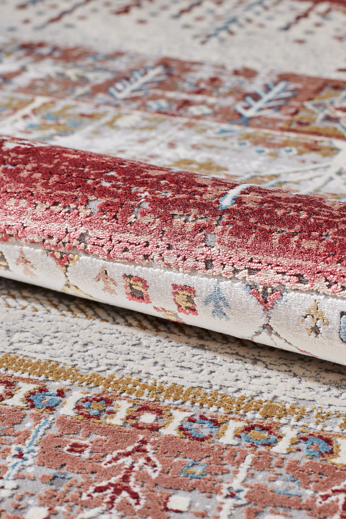 Close-up of the Vibrant Nomadica Premium Rug - M366C, highlighting its intricate traditional motifs in red, beige, blue, and gold. A folded corner reveals the rich texture and detailed woven patterns.