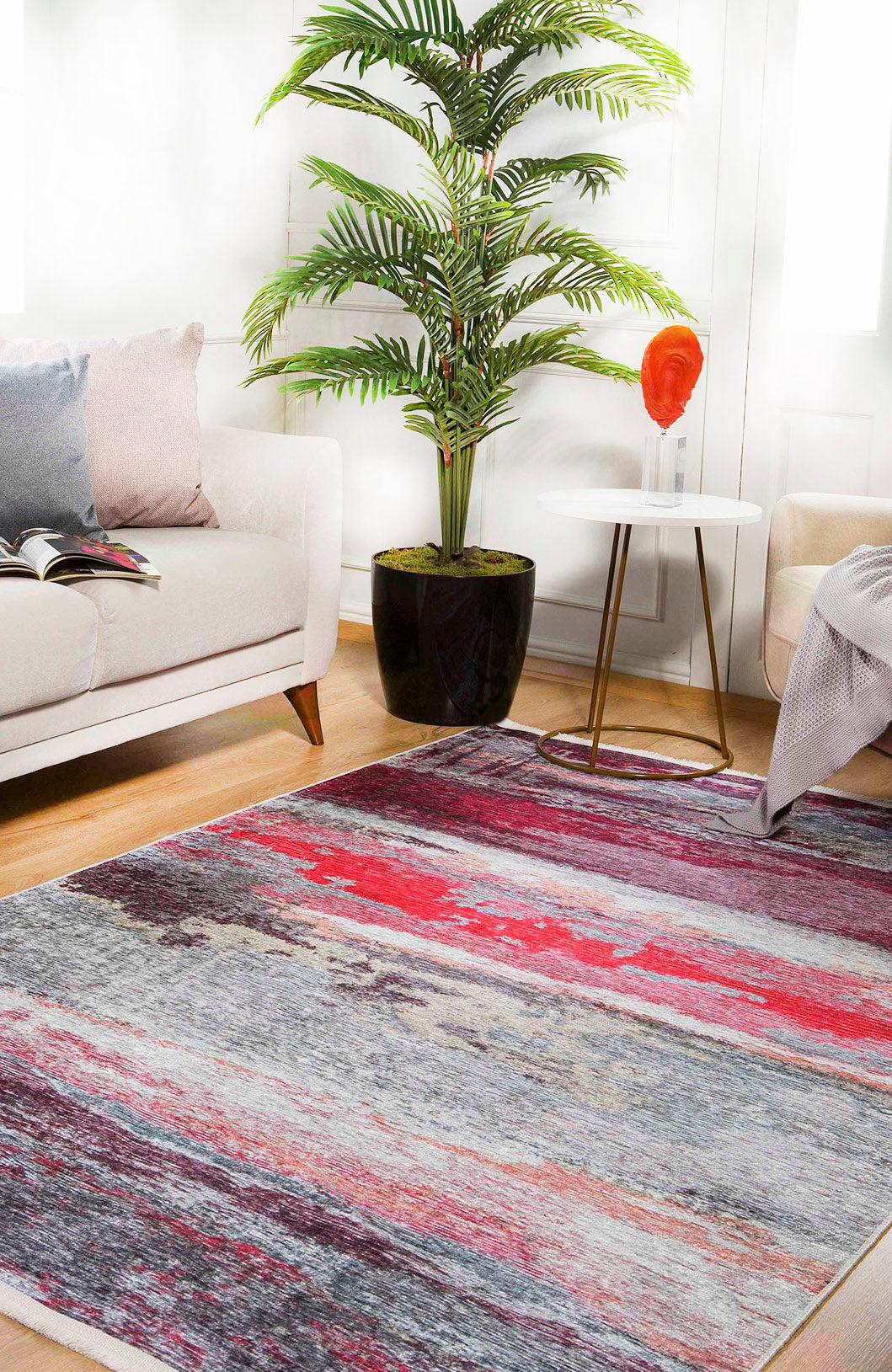 A modern living room with a large Vintage Brushed - Washable Rug - JR143 (Custom Size) in pink and red, a white sofa with pastel pillows, a potted plant, a small round side table with a red glass vase, and sunlight streaming through white doors.