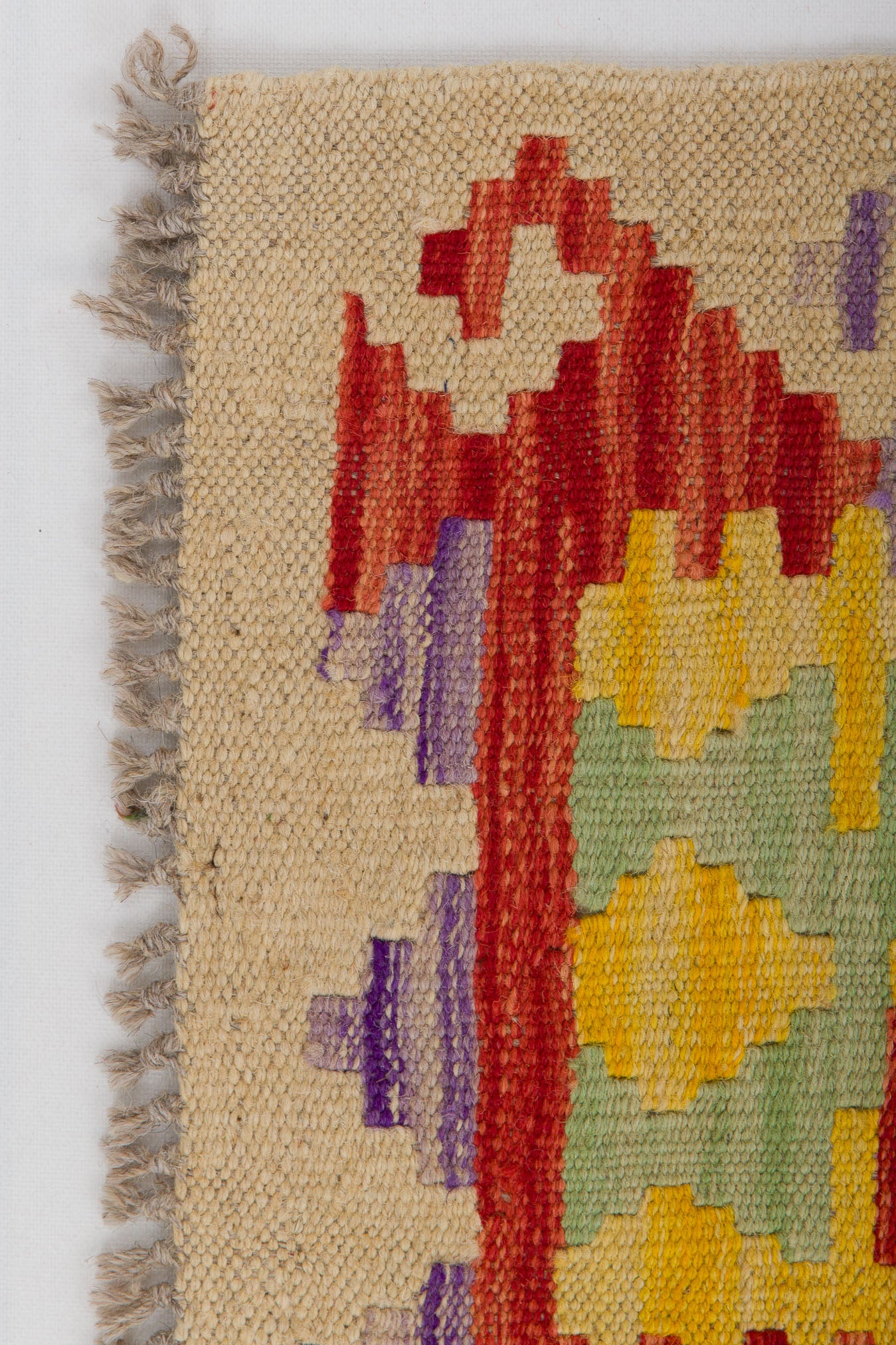A close-up of the Vintage Heritage - Handwoven Kilim flatweave rug, featuring a beige background with red, yellow, green, and purple geometric patterns and left-side fringed edges.