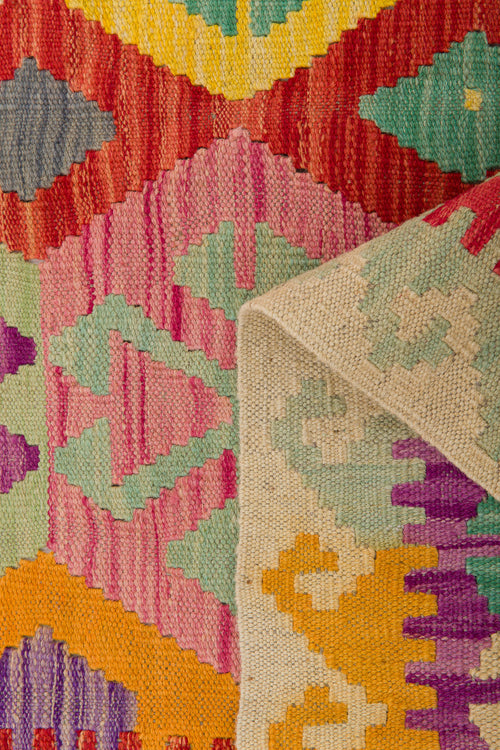 Load image 1 in gallery view. The Vintage Heritage - Handwoven Kilim is a colorful wool flatweave rug featuring geometric zigzag and triangular patterns in red, yellow, green, blue, and pink. One corner is folded over to show the backside of the fabric.