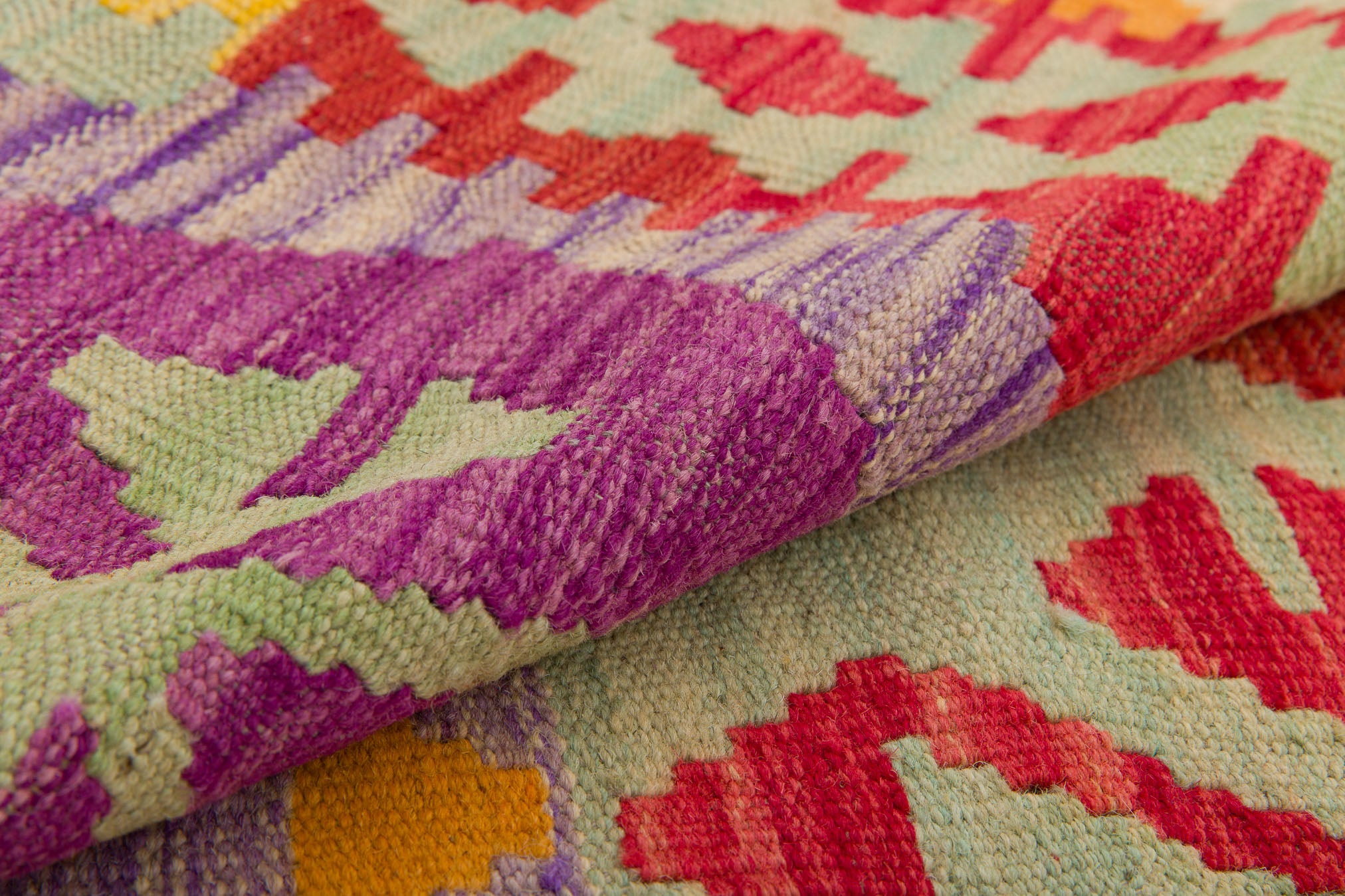 A close-up of the Vintage Heritage - Handwoven Kilim displays geometric patterns in purple, red, green, and yellow, with the folded wool rug highlighting its textured and intricate design.