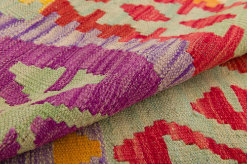 Load image 1 in gallery view. A close-up of the Vintage Heritage - Handwoven Kilim displays geometric patterns in purple, red, green, and yellow, with the folded wool rug highlighting its textured and intricate design.
