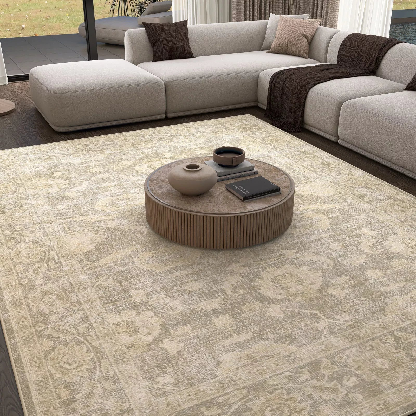 A modern living room with the Vintage Linen - Washable Rug - JRN1028 in light beige—perfect for high-traffic areas—paired with a round wooden coffee table, books, vases, and an L-shaped grey sofa with cozy throws by floor-to-ceiling windows.
