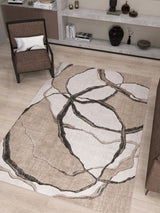 A modern living room with the Vintage Muse - Washable Rug - JRN47, a beige flat weave rug with an abstract pattern, a brown armchair with cushion, brown sofa edge, light wood floors, and shelves displaying vases and books.