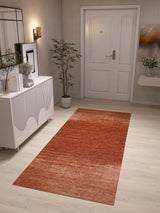 A hallway with light wood flooring features the Vintage Reverie - Washable Rug - JRN31 in a red ombré design, white cabinet, potted plant, wall art, mirror, and a white front door with a gold handle. The softly lit space is neatly decorated.