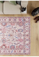 The Vivencia Authentic Washable Rug - LCC3016 (Outlet) US, featuring red, blue, and pink patterns, is displayed on a light wood floor next to a beige sofa, shoes, a bag, books, and two dark bottles.