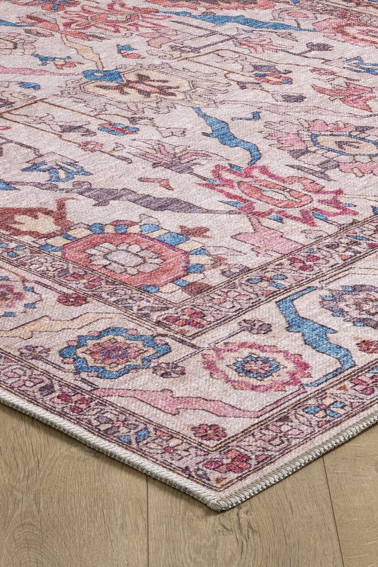 Corner of the Vivencia Authentic Washable Rug - LCC3016 (Outlet) US on a wooden floor, showcasing pink, blue, cream, and beige floral and geometric patterns with a decorative border and slightly distressed, vintage style. Durable and washable.