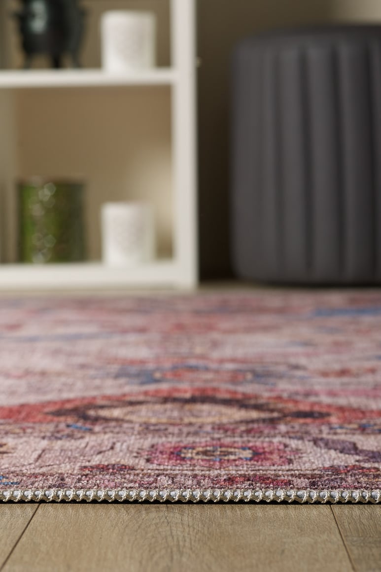 Close-up of the edge of the Vivencia Authentic Washable Rug - LCC3016 (Outlet) US on a wooden floor, with a blurred gray ottoman and white shelving unit displaying candles and decor in the background.