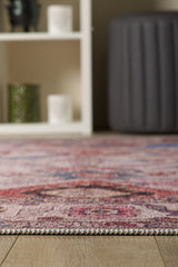 Close-up of the edge of the Vivencia Authentic Washable Rug - LCC3016 (Outlet) US on a wooden floor, with a blurred gray ottoman and white shelving unit displaying candles and decor in the background.