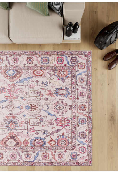 Load image 1 in gallery view. A beige sofa sits by books and a water bottle on light wood flooring near the Vivencia Authentic Washable Rug - LCC3016 (Custom Size), featuring cream, beige, pink, blue, and red patterns; a brown bag and shoes are to the right.