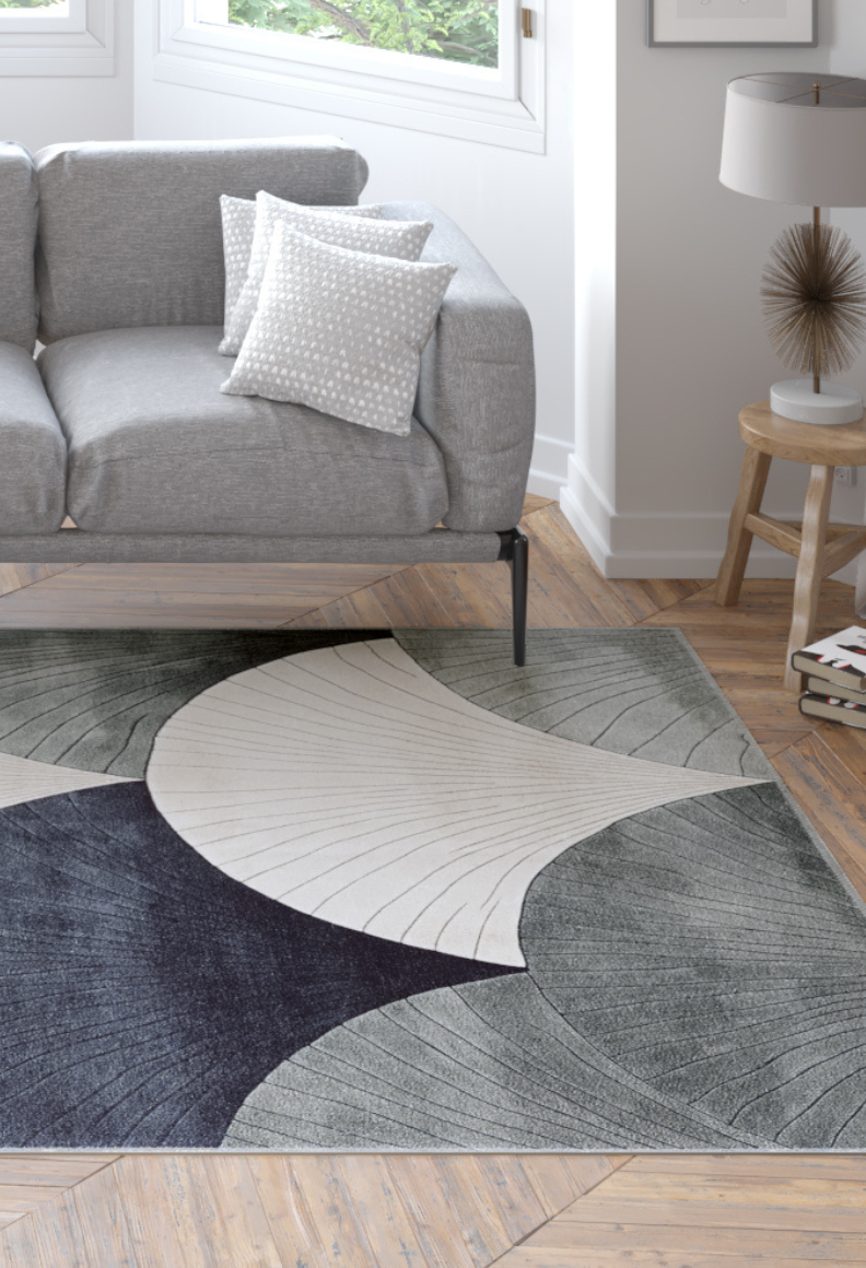 A modern living room with a gray sofa, patterned white pillows, a wooden side table and lamp, and the Vivian Modern Washable Area Rug - MDJ010 (Custom Size) in black, gray, and cream abstract shapes on a wooden floor.