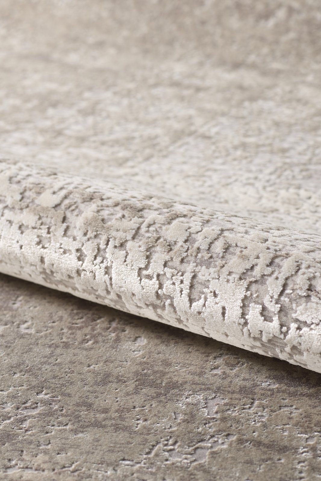 Close-up of the Vortexia Light Premium Rug - M472D in beige and light gray. This machine-made rug features a subtle abstract pattern, a soft, slightly raised surface, and is partially rolled at the edge for added style.