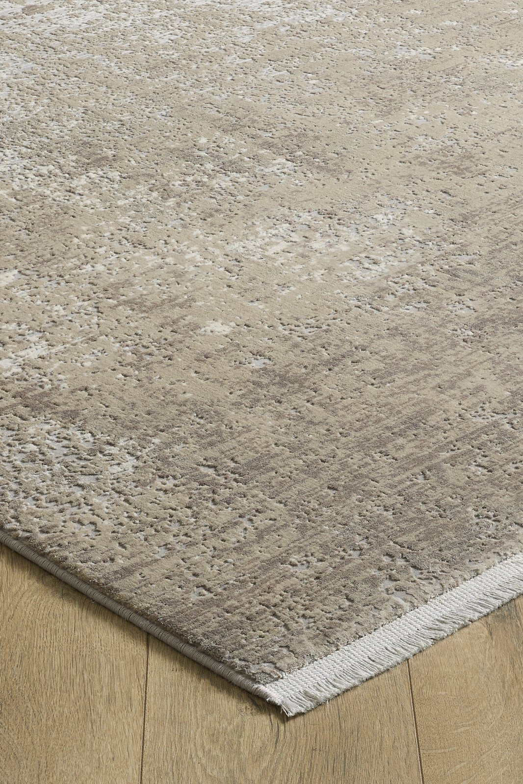 A close-up of the Vortexia Light Premium Rug - M472D, in beige and taupe with a subtle distressed texture, lies on a light wooden floor. This machine-made rug features short white fringe along its edge.