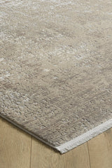 A close-up of the Vortexia Light Premium Rug - M472D, in beige and taupe with a subtle distressed texture, lies on a light wooden floor. This machine-made rug features short white fringe along its edge.