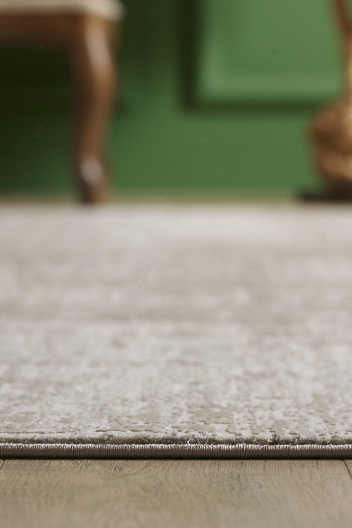Load image 1 in gallery view. A close-up of the Vortexia Light Premium Rug - M472D's edge on a wooden floor, with a blurred green wall and part of a wooden chair in the background.