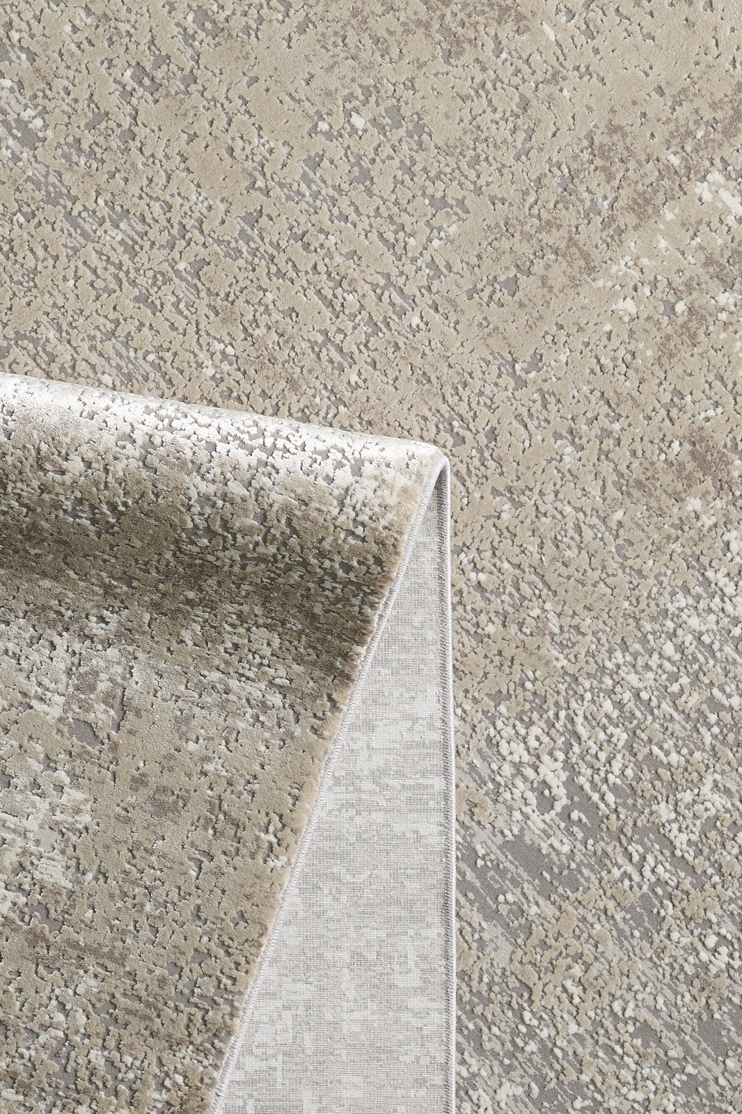 A close-up of the Vortexia Light Premium Rug - M472D in beige and gray, with a folded corner revealing the backing. This machine-made rug displays an abstract, speckled pattern in neutral tones.