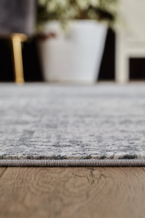 Load image 1 in gallery view. Close-up of the Vortexia Silver Premium Rug - M472A on a wooden floor, with a white planter and a chair with gold legs softly blurred in the background.