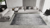 A modern living room featuring the Vortexia Silver Premium Rug - M472A, a light gray sectional with assorted cushions, a small side table, a black lounge chair, and minimalist neutral décor.