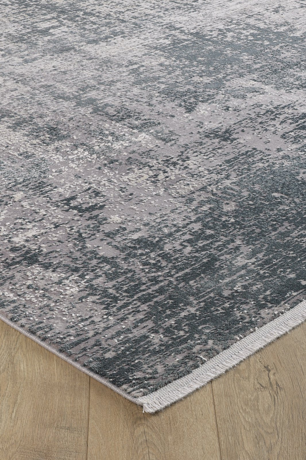 A close-up of the Vortexia Silver Premium Rug - M472A, showcasing its distressed texture, abstract gray and blue pattern, and short fringe on the edge, placed atop light hardwood flooring.