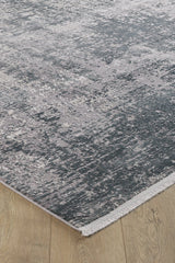 A close-up of the Vortexia Silver Premium Rug - M472A, showcasing its distressed texture, abstract gray and blue pattern, and short fringe on the edge, placed atop light hardwood flooring.