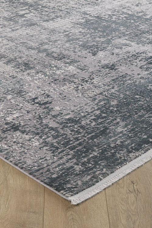 Load image 1 in gallery view. A close-up of the Vortexia Silver Premium Rug - M472A, showcasing its distressed texture, abstract gray and blue pattern, and short fringe on the edge, placed atop light hardwood flooring.