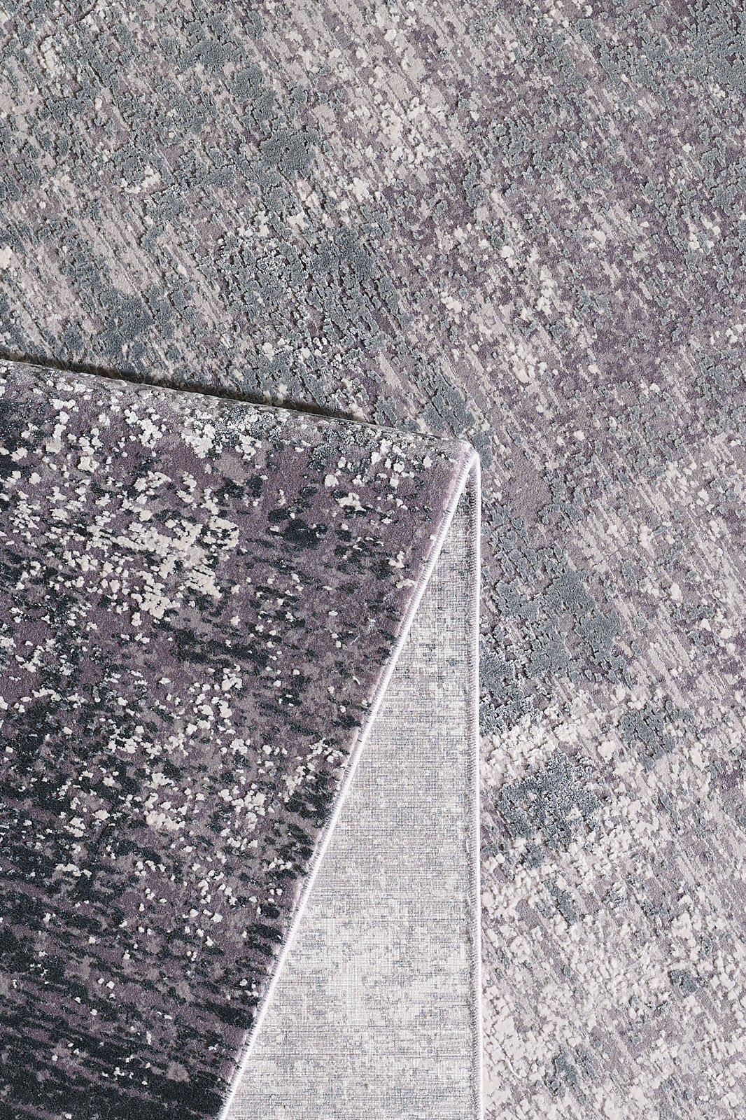 A close-up of the Vortexia Silver Premium Rug - M472A highlights its gray and purple tones, with one corner folded to reveal the lighter underside and intricate patterns on both sides.