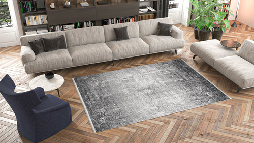 Load image 1 in gallery view. A modern living room with a long beige sofa, gray cushions, a round coffee table, blue armchair, wooden herringbone floor, bookshelves, tall green plants, and the Vortexia Silver Premium Rug - M472A featuring a distressed texture.