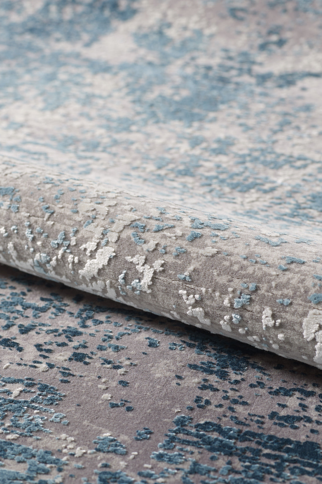 A close-up of the Vortexia Sky Premium Rug - M472B shows its soft, plush feel and abstract, cloud-like designs in blue, grey, and white with a subtle distressed texture. The contemporary machine-made rug is partially rolled or folded.