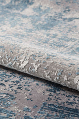 A close-up of the Vortexia Sky Premium Rug - M472B shows its soft, plush feel and abstract, cloud-like designs in blue, grey, and white with a subtle distressed texture. The contemporary machine-made rug is partially rolled or folded.