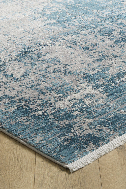 Load image 1 in gallery view. A close-up of the Vortexia Sky Premium Rug - M472B, a rectangular rug with a blue, gray, and white abstract pattern, distressed texture, and short white fringe on a light wood floor.