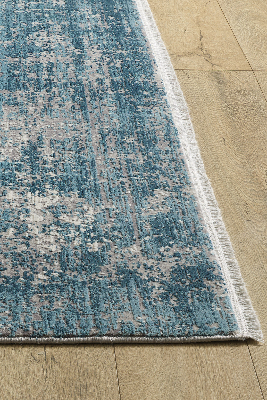 A close-up of the Vortexia Sky Premium Rug - M472B, a rectangular blue and white machine-made rug with white fringed edges and a textured abstract design, displayed on a light wood floor.