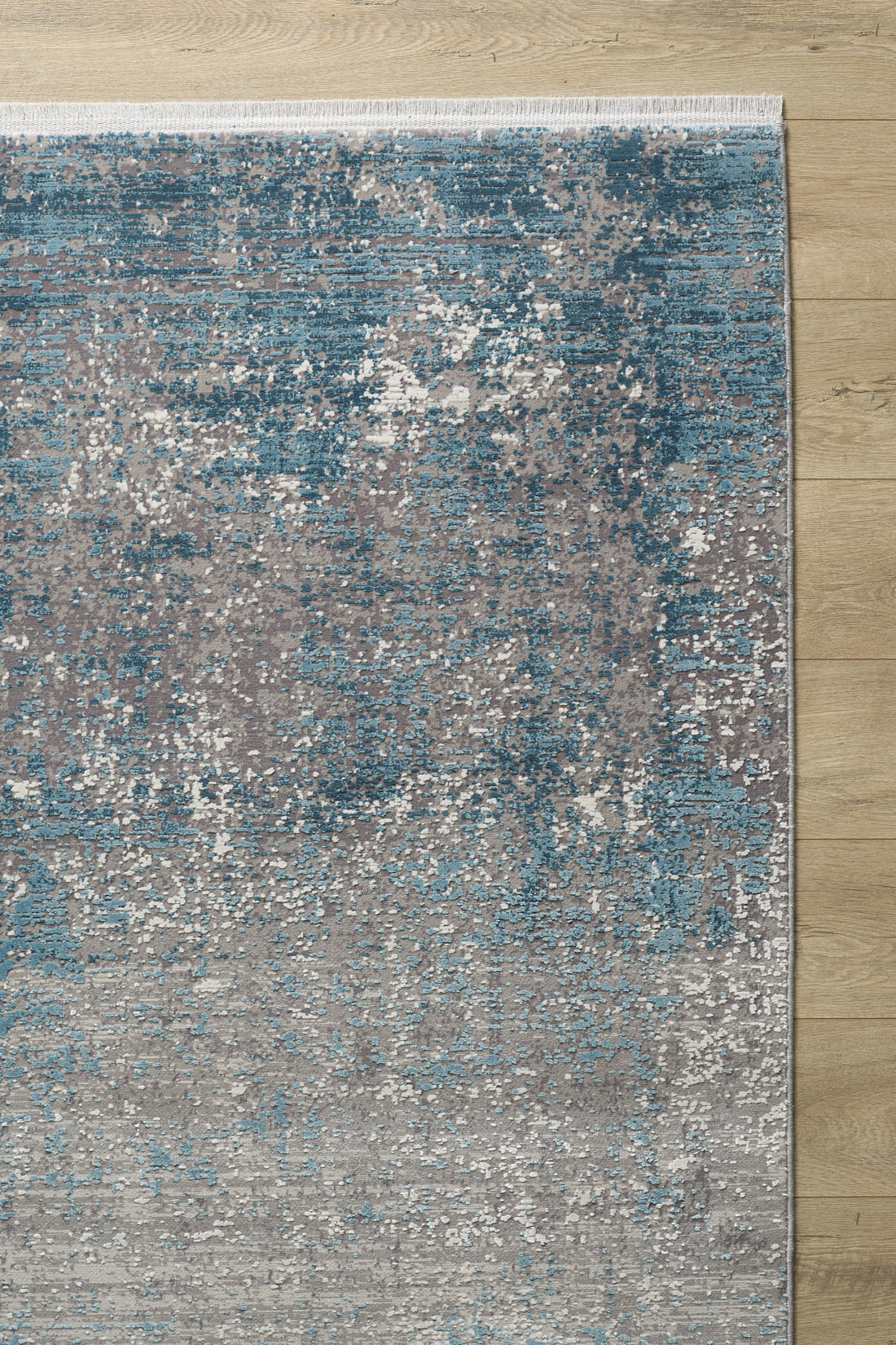 A close-up of the Vortexia Sky Premium Rug - M472B, showcasing its textured abstract pattern in blue, gray, and white on a light wooden floor. The machine-made rug has a white fringe along its top edge.