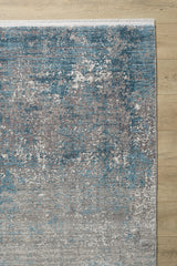 A close-up of the Vortexia Sky Premium Rug - M472B, showcasing its textured abstract pattern in blue, gray, and white on a light wooden floor. The machine-made rug has a white fringe along its top edge.