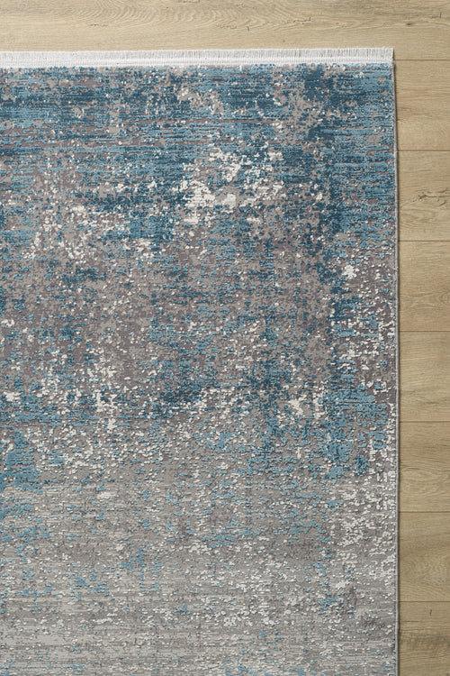 Load image 1 in gallery view. A close-up of the Vortexia Sky Premium Rug - M472B, showcasing its textured abstract pattern in blue, gray, and white on a light wooden floor. The machine-made rug has a white fringe along its top edge.