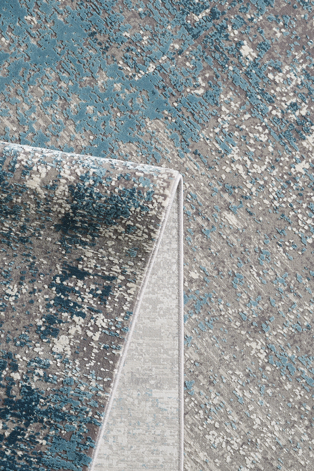 A close-up of the Vortexia Sky Premium Rug - M472B in blue and gray, featuring an abstract pattern with one corner folded to reveal the underside and showcase its textured detail.
