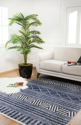 A modern living room includes a light gray sofa, the Wavy Wonders - Washable Rug - JR941 (Custom Size) in blue for high-traffic areas, a large potted plant, a white faux fur rug with magazines, crisp white walls, and a bright window.